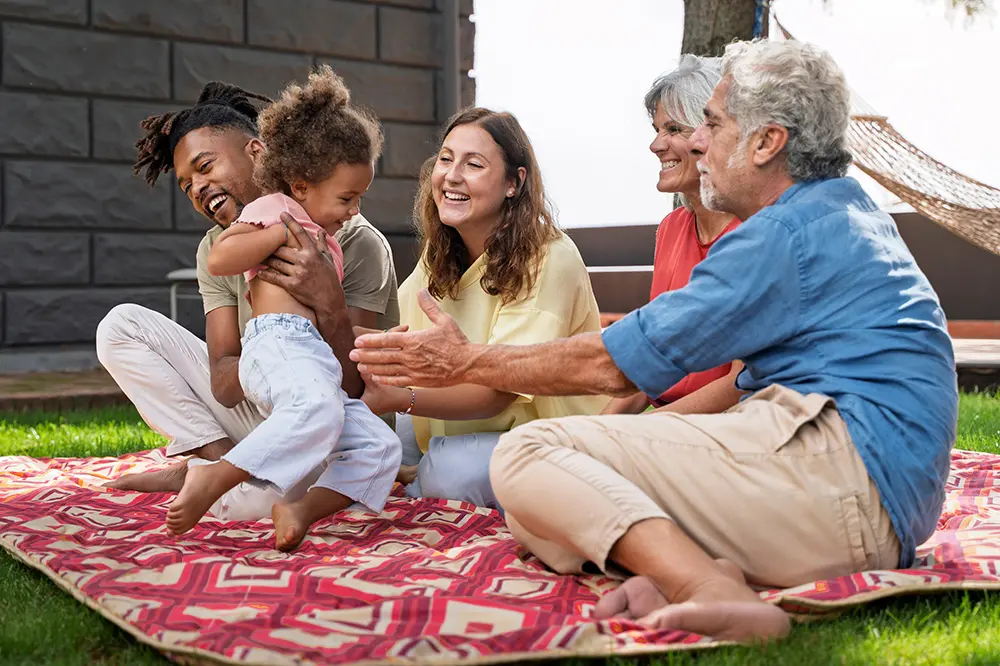 A group of five people, including a young child, sit and laugh together on a patterned blanket in a backyard. The adults smile and interact warmly as the child plays, creating a joyful, relaxed atmosphere outdoors.