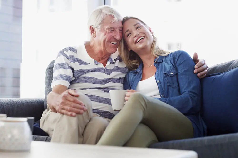 An older man and a younger woman sit closely together on a couch, smiling and laughing. The man has his arm around the woman, and she holds a mug. They appear happy and relaxed in a bright, cozy room.