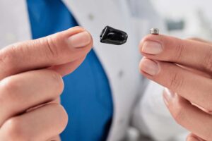 Hands holding a tiny in-canal hearing aid and a button battery, a battery life question at New River Valley Hearing & Balance in Radford, VA
