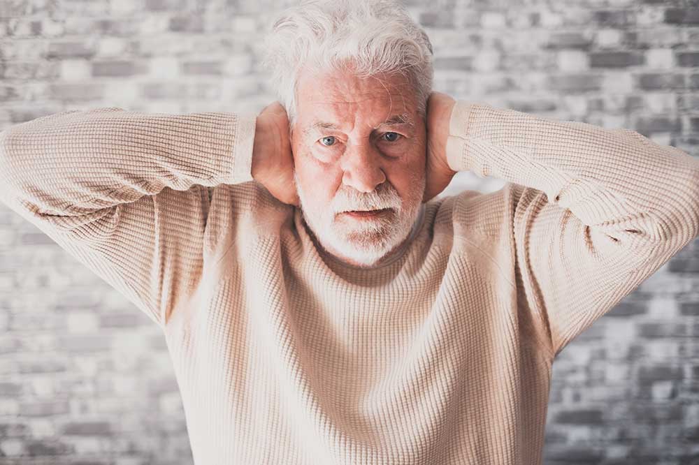 Older man with a white beard covering both ears over a muffled, clogged feeling, a concern seen at New River Valley Hearing & Balance in Radford, VA
